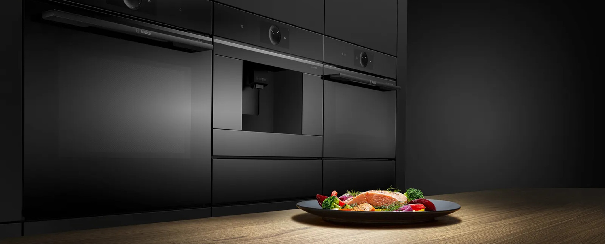 Eine köstliche Mahlzeit auf einem Teller im Hintergrund in einem Hochschrank, drei Küchengeräte von Bosch in Schwarz.