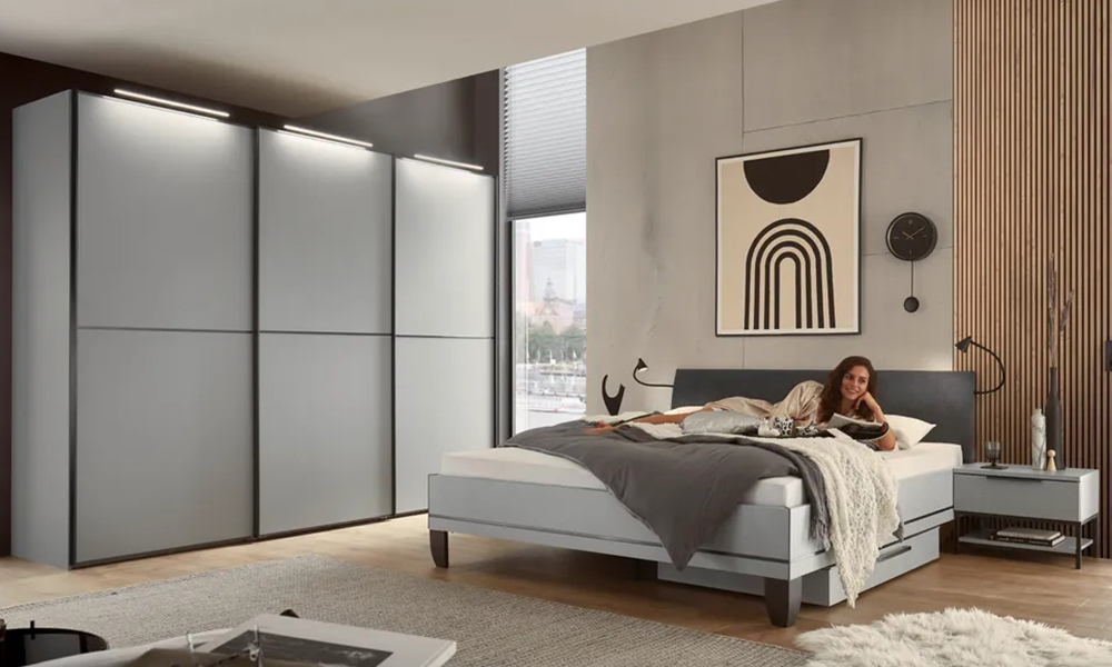 Frau liegt auf einem schlichten weißen Bett mit grauem Kopfteil, daneben steht ein 3-Türen-Kleiderschrank in der gleichen Optik.